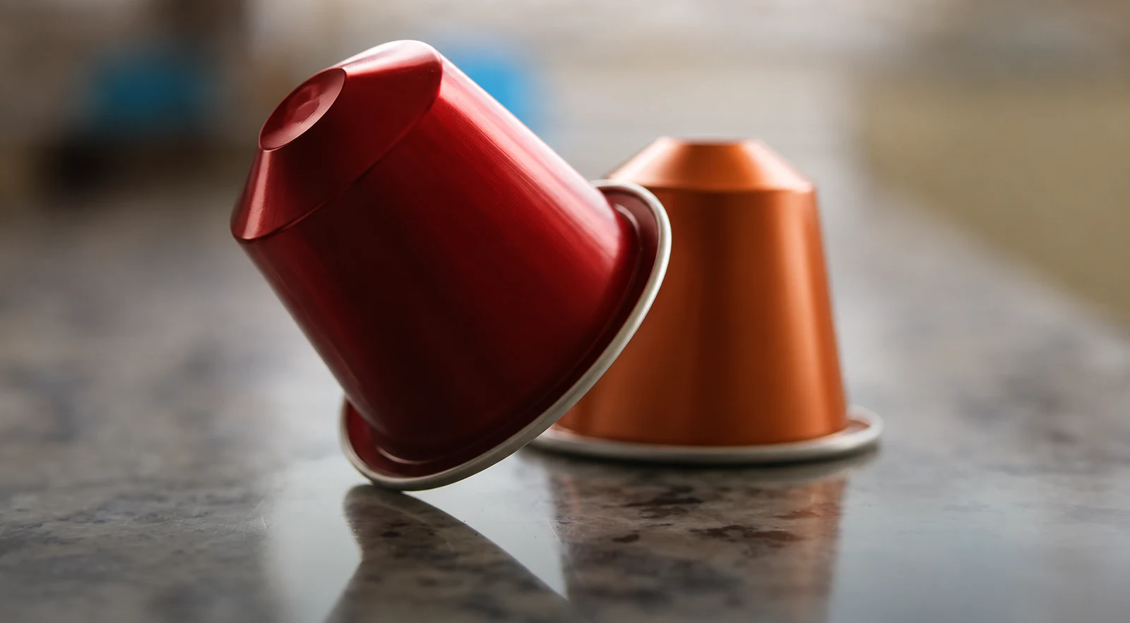 Two metallic coffee capsules arranged on a reflective stone surface.